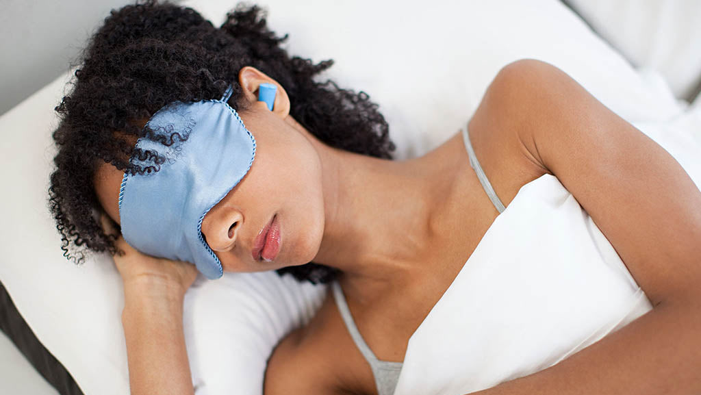 vrouw met krullend haar liggend in bed, met slaapmasker op en oordopjes in haar oren